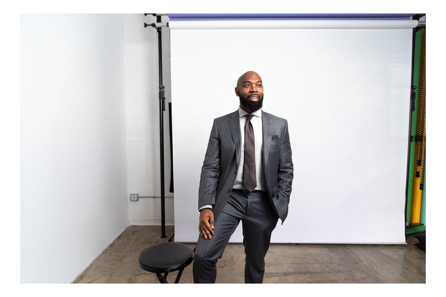 Client stands confidently in a studio portrait setup, dressed in a sharp suit, captured in a clean, minimalist environment with lighting and backdrop visible—signaling a professional, guided photo session experience.