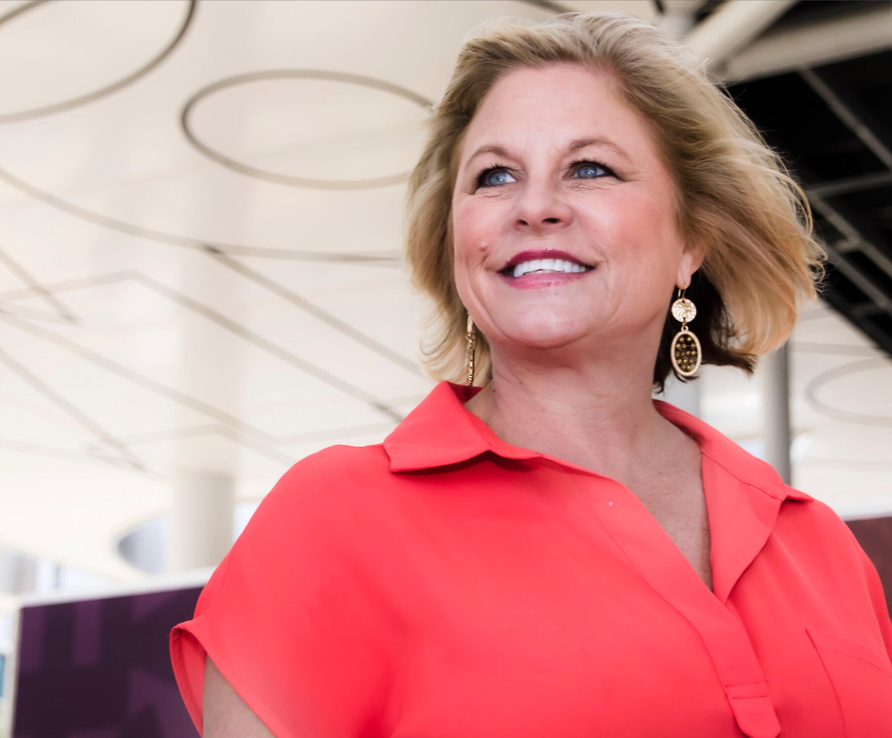 Confident woman in a bold red blouse, smiling and gazing upward in a modern architectural space — captured in soft natural light for an empowered leadership portrait.