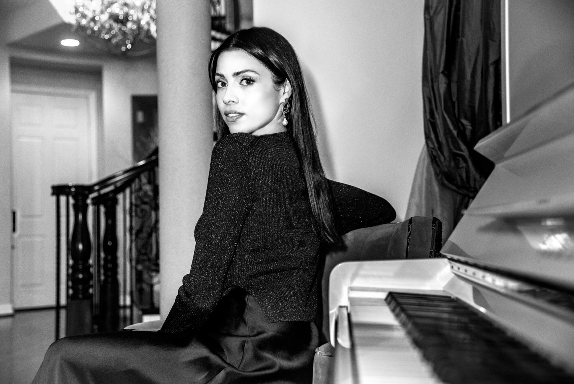 Elegant woman in black seated at a grand piano, glancing back with poised intensity — captured in high-contrast black and white for a cinematic, timeless portrait.